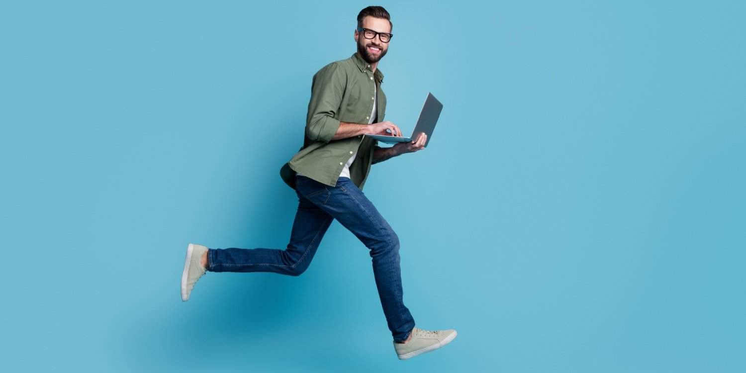 Sporty active male freelancer jumping into the air, wearing jeans, green shirt and spectacles and holding a laptop, against a blue background.