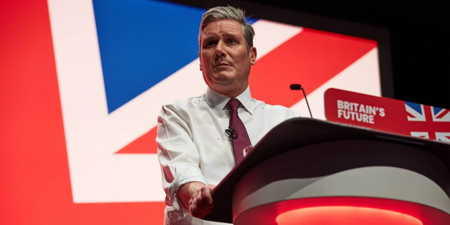 Sir Keir Starmer speaking at a conference with a white shirt and sleeves rolled up.